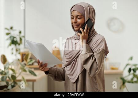 Junge lächelnde Geschäftsfrau im Hijab, die mit dem Handy spricht und dem Kunden in der häuslichen Umgebung Vertragsbedingungen vorliest Stockfoto