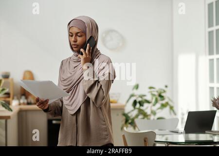 Junge Ökonomen im Hijab, die sich einen Geschäftsvertrag ansehen und mit Kollegen oder Kunden über ein Mobiltelefon in der häuslichen Umgebung sprechen Stockfoto
