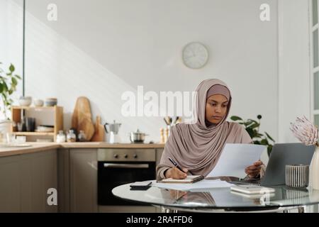 Seriöse weibliche Auditorin mit Geschäftsunterlagen, die sich die wichtigsten Punkte ansieht und sich Notizen in Copybook macht, während sie am Arbeitsplatz sitzt Stockfoto