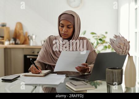 Junge, seriöse Wirtschaftsprüferin in Hijab macht sich Notizen im Copybook, während sie am Arbeitsplatz sitzt und Finanzdaten im Dokument analysiert Stockfoto