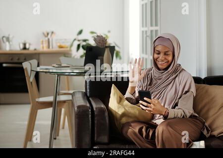 Glückliche junge muslimische Frau winkt ihrem Freund oder Ehemann auf dem Smartphone-Bildschirm die Hand, während sie zu Hause in einem Video-Chat kommuniziert Stockfoto