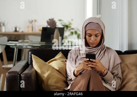 Junge, ernsthafte muslimische Frau, die auf einem schwarzen Ledersofa zwischen weichen Kissen sitzt und SMS schreibt oder Online-Videos auf einem Smartphone ansieht Stockfoto