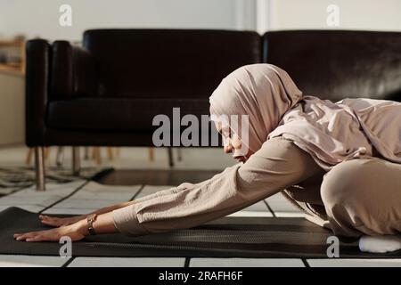 Seitenansicht einer jungen muslimischen Frau, die ihre Arme nach vorne streckt, während sie auf der Matte sitzt, ihre Beine überkreuzt und Yoga-Übungen durchführt Stockfoto