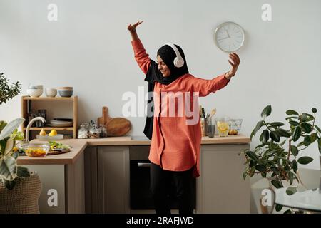 Junge, fröhliche muslimische Frau in Hijab und Kopfhörern tanzen in der Küche vor der Theke mit frischem gehacktem Gemüse in der Schüssel Stockfoto