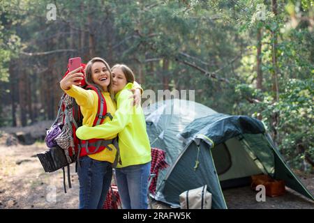 Mom mit Tochter macht ein Selfie in einem Waldlager. Sie stehen neben dem Zelt und machen ein Foto mit einem Smartphone. Mutterschaft und Vertrauen der jüngeren g Stockfoto