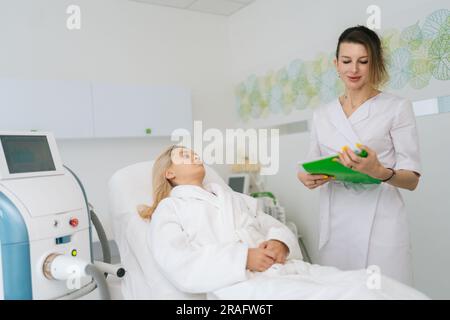 Porträt einer jungen blonden Frau im weißen Bademantel, die bei einem kosmetischen Besuch in einer bequemen Couch-Kosmetikklinik liegt und mit einem erfahrenen Arzt spricht Stockfoto