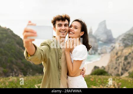 Glückliches europäisches Paar nach der Verlobung, das Selfie auf dem Handy machte, auf einer Felsklippe in der Nähe des Ozeans stand Stockfoto