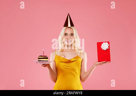 Glückliche junge Frau mit Geburtstagsmütze, Kuchen und roter Geschenkbox, Posen auf rosa Hintergrund, Studioaufnahme Stockfoto