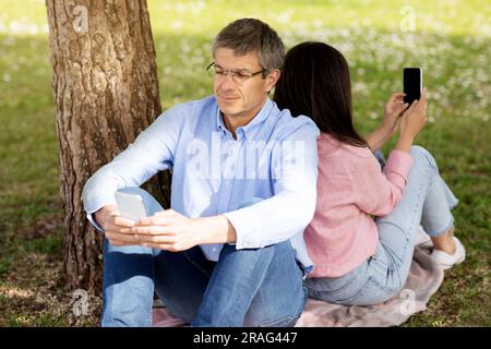 Ein Paar mittleren Alters, das sich Rücken an Rücken lehnt und sein Smartphone im Freien benutzt Stockfoto