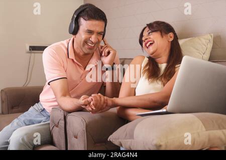 Indisches glückliches Paar essen Salat und gesunde Ernährung, sitzen auf einem Sofa mit einem Laptop Surfen online. Musik hören. Ich höre Lieder. Stockfoto