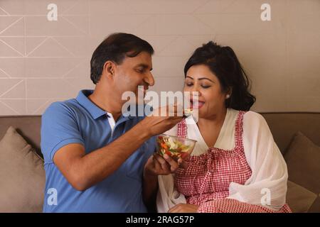 Glückliche indische gesundheitsbewusste Ehemann Ehefrau, die zusammen sitzt und eine Schüssel grünen Salat auf der Couch im Wohnzimmer isst Stockfoto