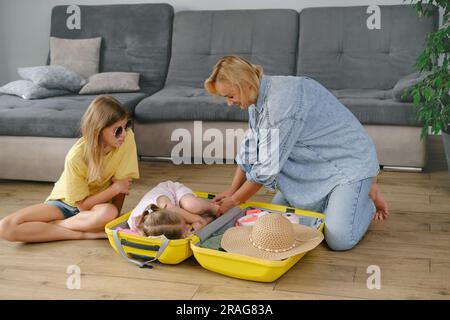 Mutter mit Töchtern, die einen gelben Koffer für die Reise packen. Die Familie hat Spaß beim Urlaub. Glückliches Kind, das im Koffer sitzt und sich auf ein vorbereitet Stockfoto