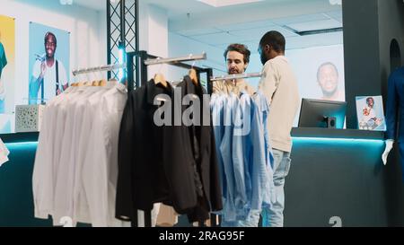 Männlicher Käufer bezahlt für trendige Kleidersammlung, kauft am POS-Terminal im Bekleidungsgeschäft. Junger Mann, der mit einem Boutique-Mitarbeiter an der Kasse spricht, kleines Unternehmen. Handgeführte Aufnahme. Stockfoto