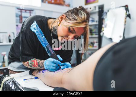 Tätowierte Tätowierungsmeisterin mit Schutzhandschuhen, die ein großes Tattoo auf einen Körper malt, mittlere Aufnahme. Hochwertiges Foto Stockfoto