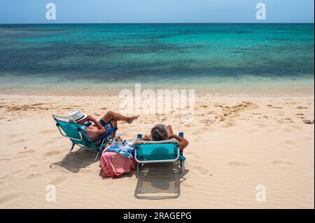 Touristen ruhen sich in den Sesseln aus und genießen das heiße Sommerwetter am kristallklaren türkisfarbenen Strand von Corralejo, bekannt als „Grandes Playas de Corralejo“ Stockfoto