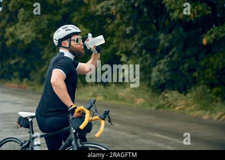 Bärtiger junger Mann, Radfahrer in Uniform, Helm und Gläser stehen auf der Straße und Trinkwasser. Erfrischung und Erholung Stockfoto