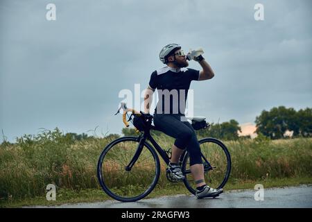 Bärtiger junger Mann, Radfahrer in Uniform, Helm und Gläser stehen auf der Straße und Trinkwasser. Bewölkter Abend Stockfoto
