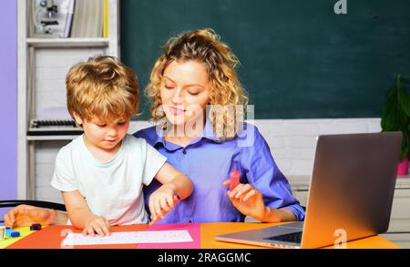 Eine Grundschullehrerin, die dem kleinen Jungen beim Schreibunterricht in der Schule hilft. Junge Lehrerin, die das Kind auf die Schulprüfung vorbereitet Stockfoto