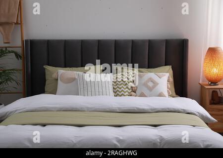 Komfortables Bett mit Kissen, Lampe auf einem Nachttisch aus Holz im Zimmer. Stilvolle Innenausstattung Stockfoto