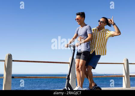 Glückliches birassisches, schwules Paar, das Spaß hat, zusammen auf dem Roller auf der Promenade auf dem Meer zu fahren, Kopierraum Stockfoto