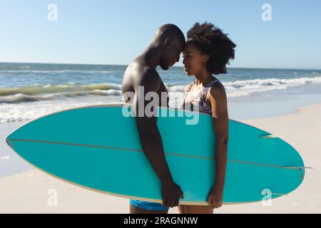 Glückliches afroamerikanisches Paar mit Surfbrett, das am sonnigen Strand steht und Köpfe berührt Stockfoto