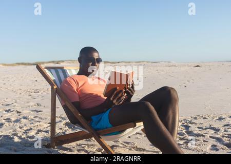 Ein glücklicher afroamerikanischer Mann mit Sonnenbrille, der im Liegestuhl sitzt und am sonnigen Strand ein Buch liest Stockfoto