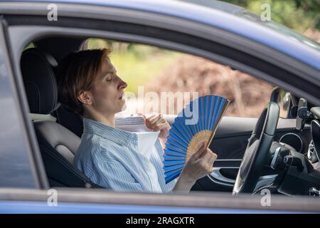 Eine müde, erschöpfte Frau mittleren Alters, die mit einem blauen Fächer wedelt, leidet an Steifigkeit im Auto. Stockfoto