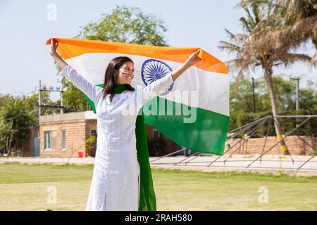 Glückliche junge Frau in traditionellem weißen Kleid, die im Park die indische Flagge im Freien weben, um den Unabhängigkeitstag oder den Republikstag zu feiern. Stockfoto