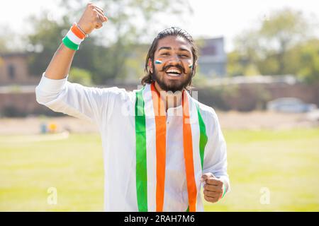 Porträt eines fröhlichen jungen Mannes, der traditionelle weiße Kurta und dreifarbige Duppata trägt, mit einem Gesicht gemalt, das die indische Sportmannschaft anfeuert, feiert Stockfoto