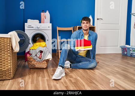 Junger lateinamerikanischer Mann, der saubere Wäsche entspannt hält, mit ernstem Gesichtsausdruck. Einfaches und natürliches Betrachten in die Kamera. Stockfoto