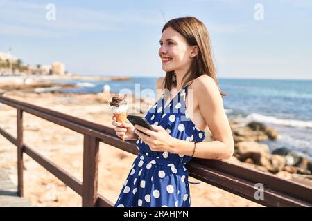 Junge blonde Touristin, die Eis am Meer mit einem Smartphone hält Stockfoto