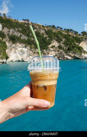 Handgehaltene Plastiktasse Eiskaffee im Sommerurlaub mit Meer im Hintergrund Stockfoto