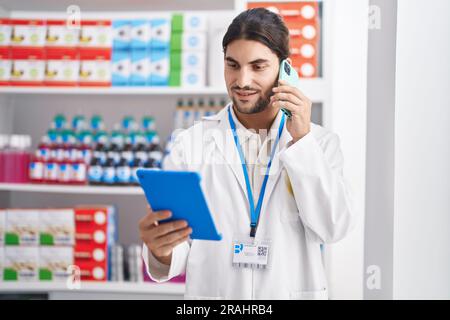 Junger hispanischer Apotheker mit Touchpad, der in der Apotheke auf dem Smartphone spricht Stockfoto