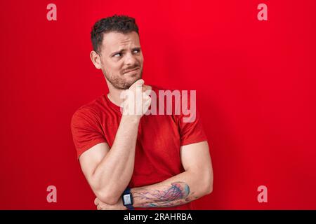 Junger lateinamerikanischer Mann, der über rotem Hintergrund steht und sich Sorgen um eine Frage macht, besorgt und nervös mit der Hand am Kinn Stockfoto