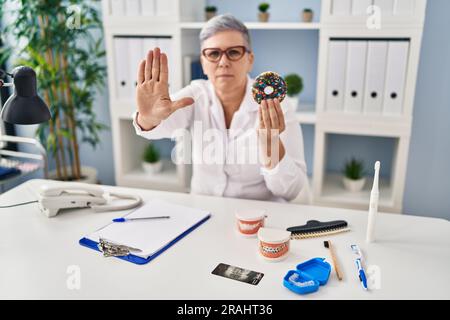 Zahnärztin mittleren Alters, die einen Schoko-Donut in der Hand hält, lächelt mit Händen auf der Brust, Augen geschlossen mit dankbarer Geste im Gesicht. Gesundheitskonzept. Stockfoto