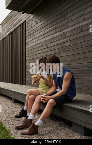 Lächelnde brünette Frau im Sommerkostüm und Stiefel mit frischem Brötchen im Handwerkspaket und neben einem stilvollen Freund mit Kaffee zum Mitnehmen in der Nähe von Holz Stockfoto