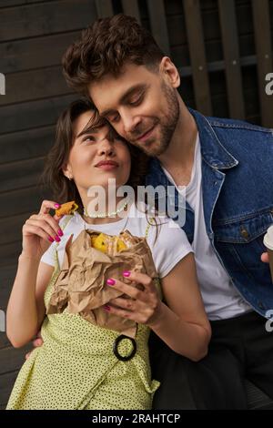 Porträt einer freudigen brünetten Frau in einem Sonnenkleid, die einen frischen Brötchen im Handwerkspaket hält und einen stilvollen Freund in Jeansweste sieht, während sie neben Woo sitzt Stockfoto