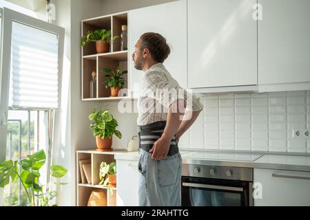 Mann mit Rückenstützgurt am unteren Rücken zur Behandlung von Hernien in der Küche mit Blick auf das Fenster. Stockfoto