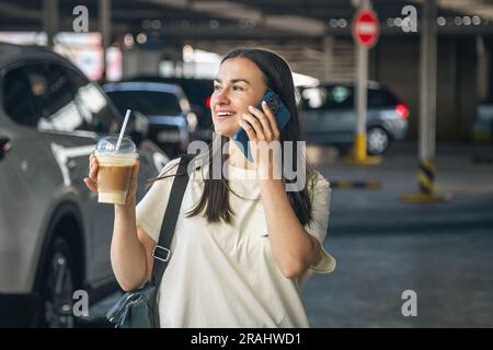 Eine junge Frau mit Kaffee am Telefon auf dem Parkplatz. Stockfoto