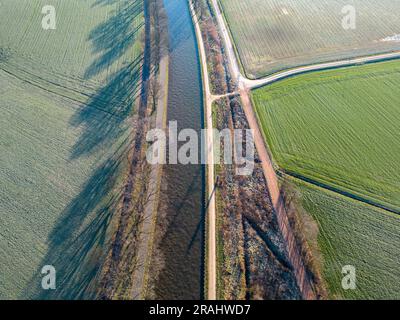 Erleben Sie die ruhige Schönheit des Winters, während die Kamera über einem ruhigen Fluss oder Kanal inmitten der gefrorenen Farmfelder schwingt. Diese fesselnde Luftaufnahme zeigt die bezaubernde Stille und Reinheit der Saison. Die eisige Wasserstraße schlängelt sich anmutig durch die schneebedeckten Felder und schafft einen faszinierenden Kontrast zwischen dem kühlen Blau des Flusses und dem unberührten Weiß der umliegenden Landschaft. Erleben Sie die Symphonie der Natur und die Berührung des Winters, während die Luftperspektive die atemberaubende Ruhe und Einsamkeit enthüllt, die die Landschaft bedeckt. Erleben Sie die Ruhe und Stockfoto