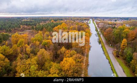 Tauchen Sie ein in den lebendigen Herbstteppich, während die Kamera einen bezaubernden Blick auf einen Fluss oder Kanal, der durch den Wald fließt, einfängt mit farbenfrohen Herbstlaub vor einem dramatischen bewölkten Himmel einfängt. Die Natur wird lebendig, wenn die Blätter sich in ein Kaleidoskop aus warmen Farbtönen verwandeln und die Landschaft mit Rot-, Orange- und Goldtönen bemalen. Die ruhige Wasserstraße schlängelt sich durch die malerischen Wälder und spiegelt die fesselnden Farben oben wider. Der dramatisch bewölkte Himmel sorgt für Tiefe und Kontrast und schafft eine stimmungsvolle Kulisse, die die Schönheit des Herbstfeuers verstärkt Stockfoto