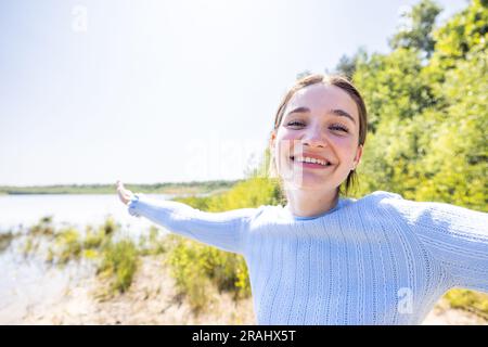 Strahlende Freiheit: Eine fröhliche Frau, die die Schönheit der Natur an einem Waldsee begrüßt, eine freie, glückliche Frau, die die Natur an einem Waldsee genießt. Beauty Girl Im Freien. Freiheitskonzept. Wunderschöne attraktive Girl Over Sky und Sun. Sonnenbalken. Vergnügen. Hochwertige Fotos, erleben Sie die reine Essenz von Glück und Freiheit, während eine fesselnde Frau freudig die Schönheit der Natur an einem ruhigen Waldsee begrüßt. Dieses sorgenfreie und attraktive Mädchen sonnt sich im Sonnenschein, dessen Strahlen eine warme und strahlende Aura ausstrahlen. Vor dem Hintergrund des weiten Himmels schwelgt sie in der Freiheit und Ruhe, die die Natur schafft Stockfoto