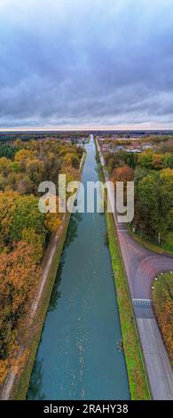 Tauchen Sie ein in den lebendigen Herbstteppich, während die Kamera einen bezaubernden Blick auf einen Fluss oder Kanal, der durch den Wald fließt, einfängt mit farbenfrohen Herbstlaub vor einem dramatischen bewölkten Himmel einfängt. Die Natur wird lebendig, wenn die Blätter sich in ein Kaleidoskop aus warmen Farbtönen verwandeln und die Landschaft mit Rot-, Orange- und Goldtönen bemalen. Die ruhige Wasserstraße schlängelt sich durch die malerischen Wälder und spiegelt die fesselnden Farben oben wider. Der dramatisch bewölkte Himmel sorgt für Tiefe und Kontrast und schafft eine stimmungsvolle Kulisse, die die Schönheit des Herbstfeuers verstärkt Stockfoto
