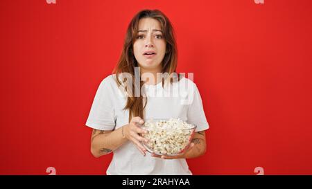 Junge, wunderschöne hispanische Frau, die Horrorfilme sieht und Popcorn auf isoliertem roten Hintergrund isst Stockfoto