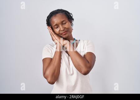 Afrikanische Frau mit Dreadlocks, die auf weißem Hintergrund steht, schläft müde, träumt und posiert mit den Händen zusammen, während sie mit geschlossenen Augen lächelt. Stockfoto