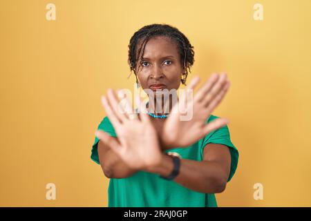 Afrikanische Frau mit Dreadlocks, die auf gelbem Hintergrund steht, mit einem Rejektionsausdruck, der Arme und Handflächen kreuzt und negative Zeichen macht, wütendes Gesicht Stockfoto
