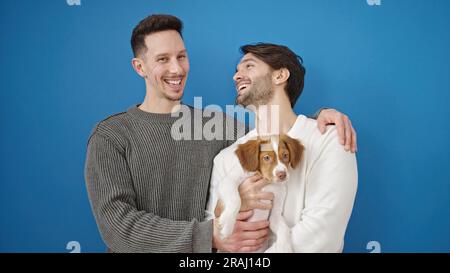 Zwei Männer, die selbstbewusst lächeln und den Hund umarmen, vor einem isolierten blauen Hintergrund Stockfoto