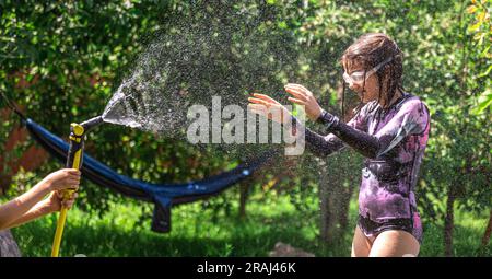 Lustiges kleines Mädchen, das im sonnigen Garten mit Gartenschlauch spielt. Stockfoto