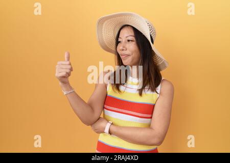 mittelalterliche chinesin mit Sommerhut auf gelbem Hintergrund, die stolz aussieht und mit Daumen-hoch-Geste zur Seite lächelt Stockfoto