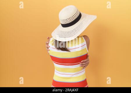 mittelalterliche chinesin mit Sommerhut auf gelbem Hintergrund, die sich glücklich und positiv von hinten umarmt. Selbstliebe und Selbstfürsorge Stockfoto